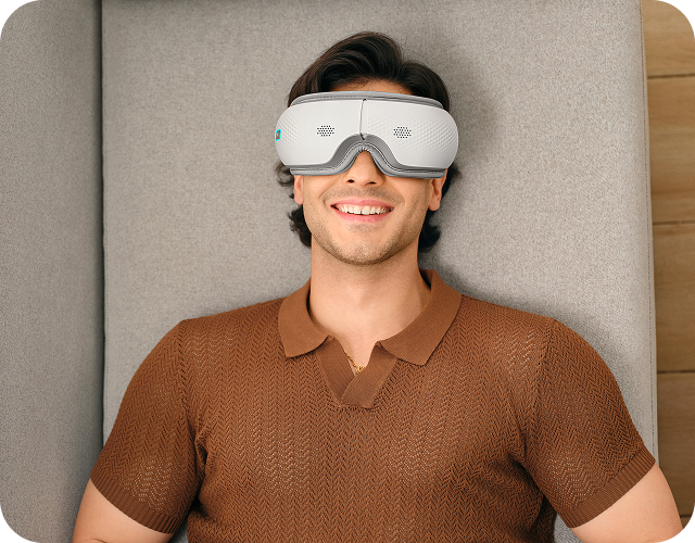 Uśmiechnięty mężczyzna siedzi w fotelu, używając białego masażera oczu SKG ES300 z niebieskimi elementami