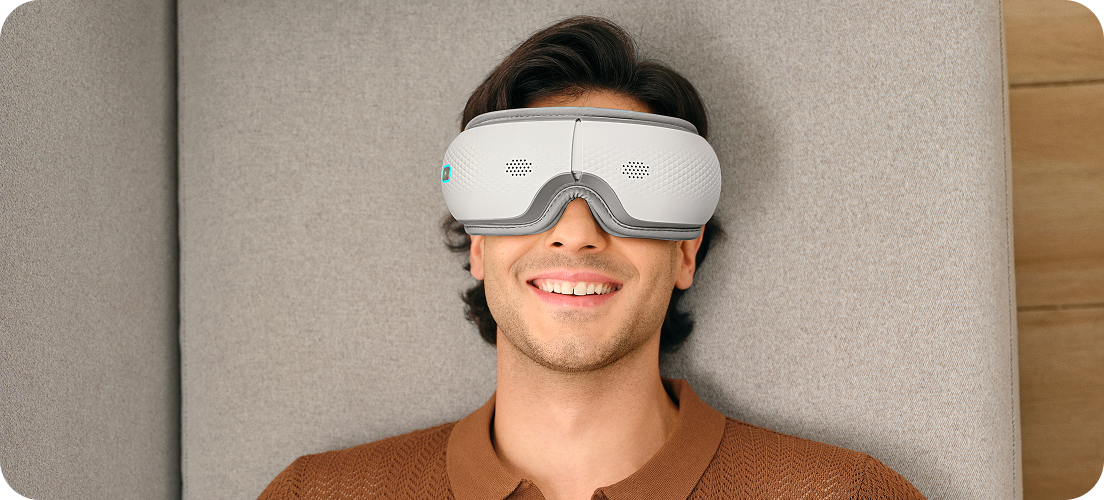 Uśmiechnięty mężczyzna siedzi w fotelu, używając białego masażera oczu SKG ES300 z niebieskimi elementami