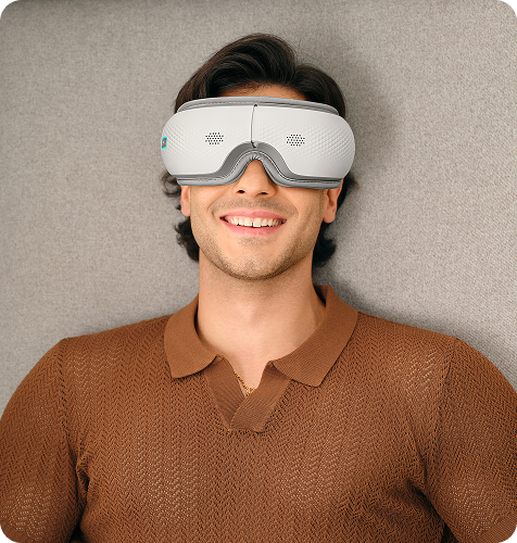 Uśmiechnięty mężczyzna siedzi w fotelu, używając białego masażera oczu SKG ES300 z niebieskimi elementami