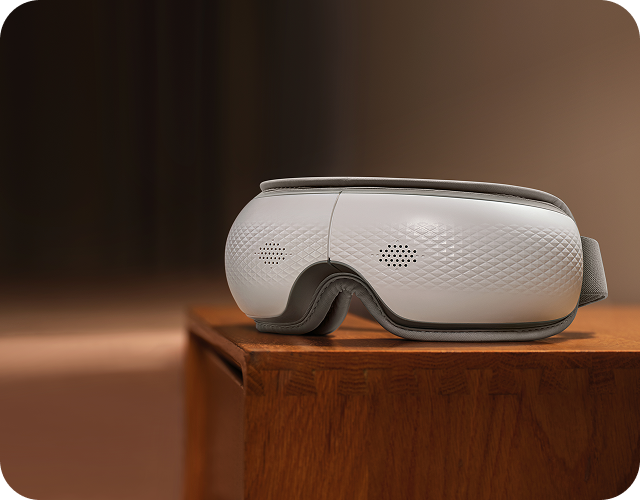 Masażer oczu SKG ES300 w kolorze biało-szarym, leżący na drewnianej komodzie w ciepłym świetle