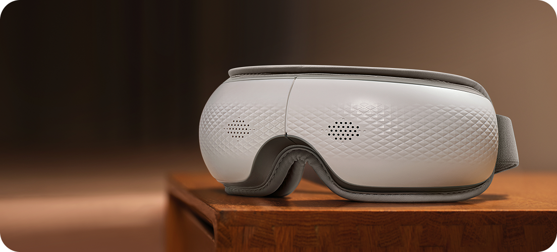 Masażer oczu SKG ES300 w kolorze biało-szarym, leżący na drewnianej komodzie w ciepłym świetle