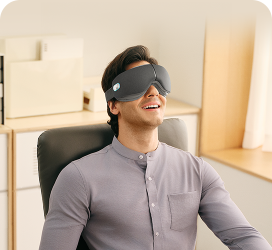 Mężczyzna siedzący w fotelu biurowym, używając szarego masażera oczu SKG ES500 podczas przerwy w pracy