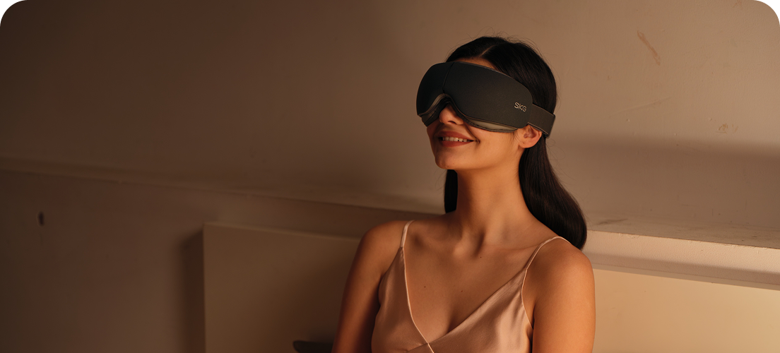 Kobieta siedząca w łóżku używa szarego masażera oczu SKG ES500, relaksując się w zaciemnionej sypialni