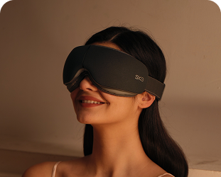 Kobieta siedząca w łóżku używa szarego masażera oczu SKG ES500, relaksując się w zaciemnionej sypialni