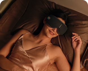 Kobieta leżąca w łóżku używa szarego masażera oczu SKG ES500, uśmiechając się podczas relaksu