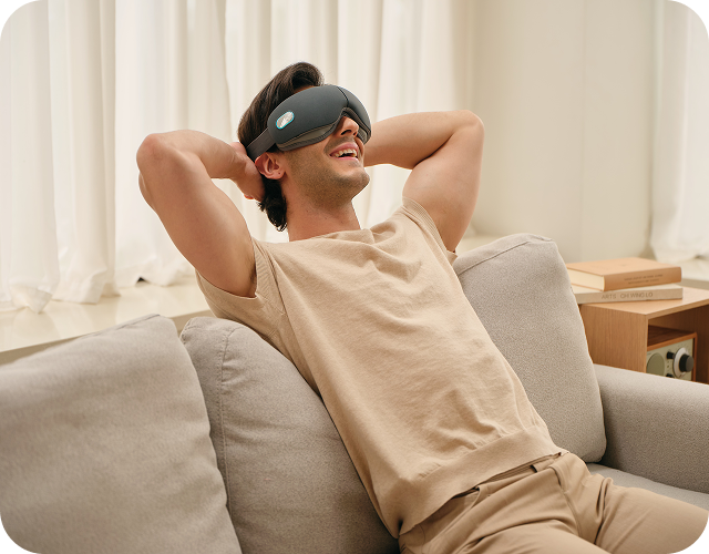 Mężczyzna uśmiecha się, relaksując się na kanapie z rękami za głową, używając szarego masażera oczu SKG ES500