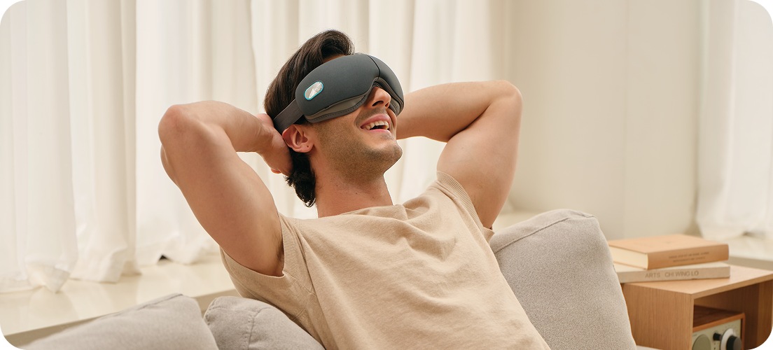 Mężczyzna uśmiecha się, relaksując się na kanapie z rękami za głową, używając szarego masażera oczu SKG ES500