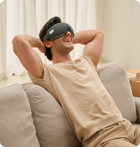 Mężczyzna uśmiecha się, relaksując się na kanapie z rękami za głową, używając szarego masażera oczu SKG ES500