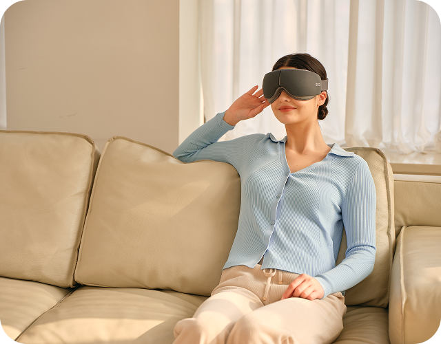 Kobieta siedzi na kanapie, używając szarego masażera oczu SKG ES500, z ręką na skroni