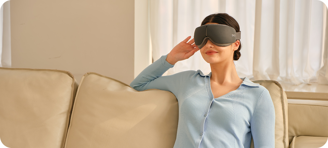 Kobieta siedzi na kanapie, używając szarego masażera oczu SKG ES500, z ręką na skroni