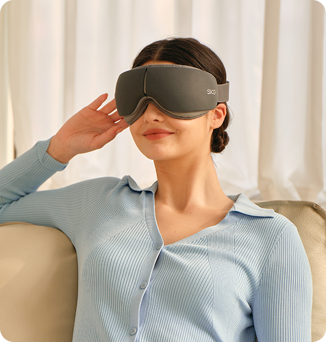 Kobieta siedzi na kanapie, używając szarego masażera oczu SKG ES500, z ręką na skroni