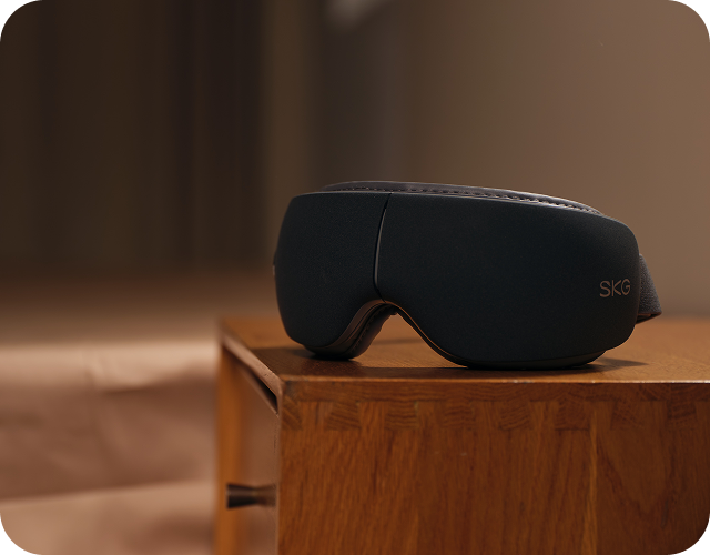 Masażer oczu SKG ES500 w kolorze szarym, leżący na drewnianej szafce nocnej w ciepłym oświetleniu