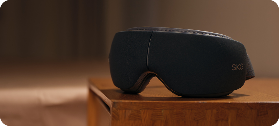 Masażer oczu SKG ES500 w kolorze szarym, leżący na drewnianej szafce nocnej w ciepłym oświetleniu