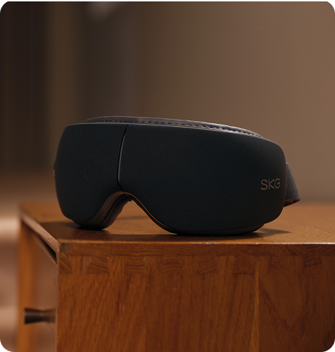 Masażer oczu SKG ES500 w kolorze szarym, leżący na drewnianej szafce nocnej w ciepłym oświetleniu