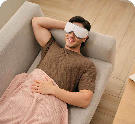 Mężczyzna leży na kanapie pod kocem, używając białego masażera oczu SKG ES500 podczas relaksu