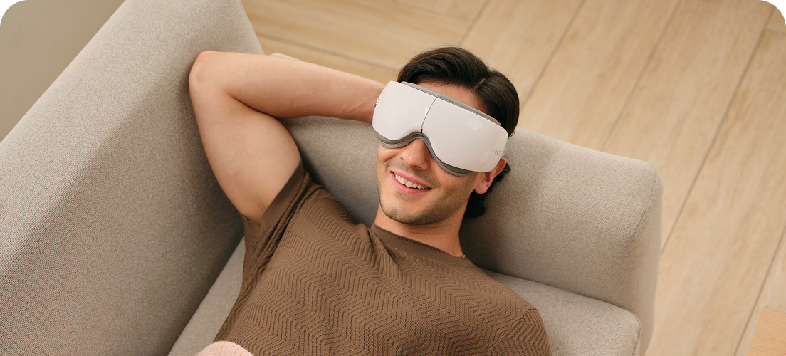 Mężczyzna leży na kanapie pod kocem, używając białego masażera oczu SKG ES500 podczas relaksu