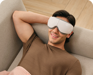 Mężczyzna leży na kanapie pod kocem, używając białego masażera oczu SKG ES500 podczas relaksu