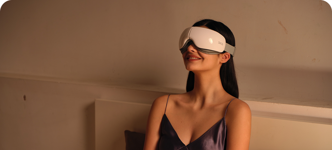 Kobieta siedząca w łóżku używa białego masażera oczu SKG ES500