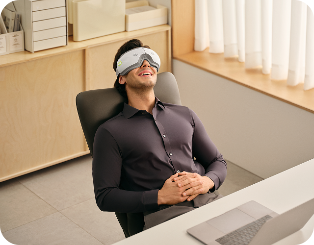 Mężczyzna siedzi w fotelu biurowym, używając białego masażera oczu SKG ES500 podczas przerwy od pracy