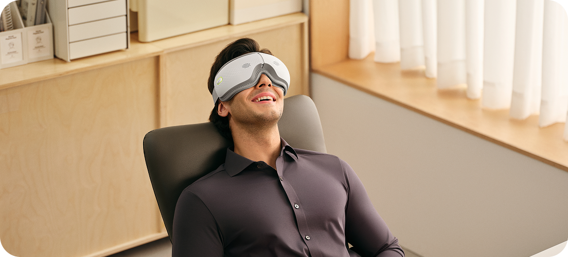 Mężczyzna siedzi w fotelu biurowym, używając białego masażera oczu SKG ES500 podczas przerwy od pracy