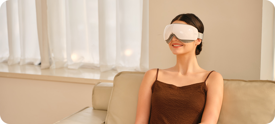 Kobieta w brązowej sukience siedzi na kanapie, używając białego masażera oczu SKG ES500 w jasnym pokoju