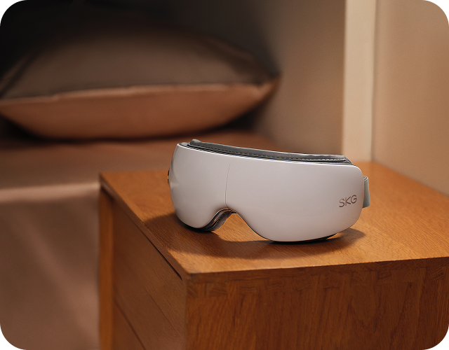 Masażer oczu SKG ES500 w kolorze białym, leżący na drewnianej szafce nocnej w sypialni