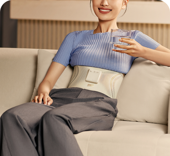 Kobieta uśmiecha się, siedząc na kanapie, używając pasa treningowego SKG GS300 i trzymając w ręce szklankę wody