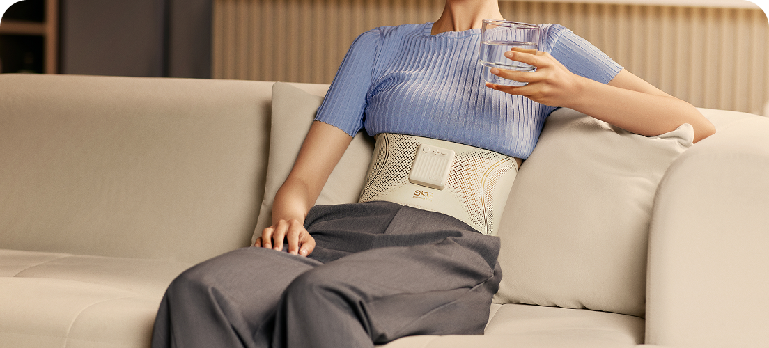 Kobieta uśmiecha się, siedząc na kanapie, używając pasa treningowego SKG GS300 i trzymając w ręce szklankę wody