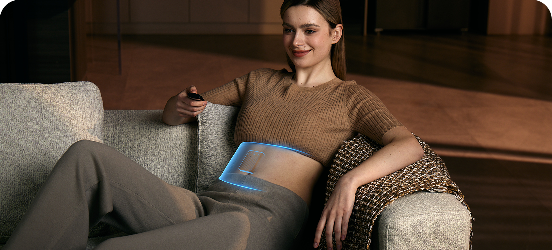 Kobieta relaksuje się na kanapie, używając pasa treningowego SKG GS300 z niebieską wizualizacją działania w okolicy brzucha