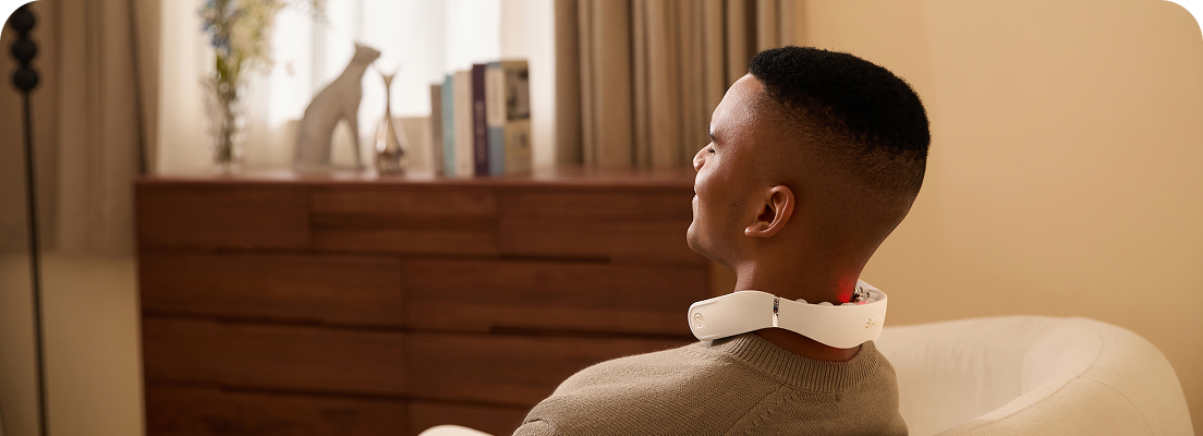 Mężczyzna relaksuje się w fotelu, używając masażera szyi SKG GS500-N ze świecącą czerwoną diodą na karku