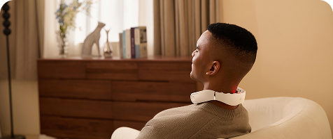 Mężczyzna relaksuje się w fotelu, używając masażera szyi SKG GS500-N ze świecącą czerwoną diodą na karku