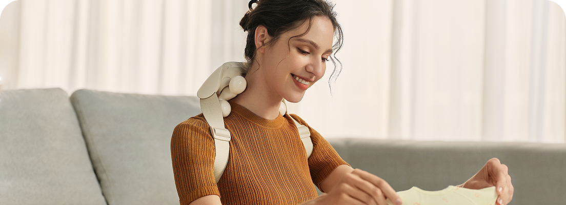 Kobieta uśmiecha się, składając ubrania dla dziecka, mając na sobie masażer karku SKG HS500-2