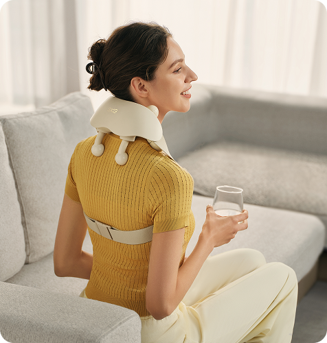 Kobieta siedzi na kanapie z masażerem karku SKG HS500-2 i paskiem mocującym, trzymając w ręce szklankę