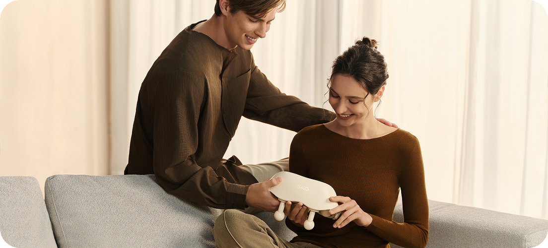 Para na kanapie ogląda masażer karku SKG HS500-2 w kolorze beżowym, trzymając go razem w rękach