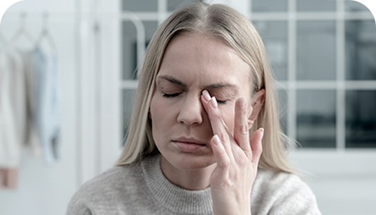 Kobieta z zamkniętymi oczami i zmartwioną miną dotyka palcami skroni, prawdopodobnie odczuwając ból lub zmęczenie.