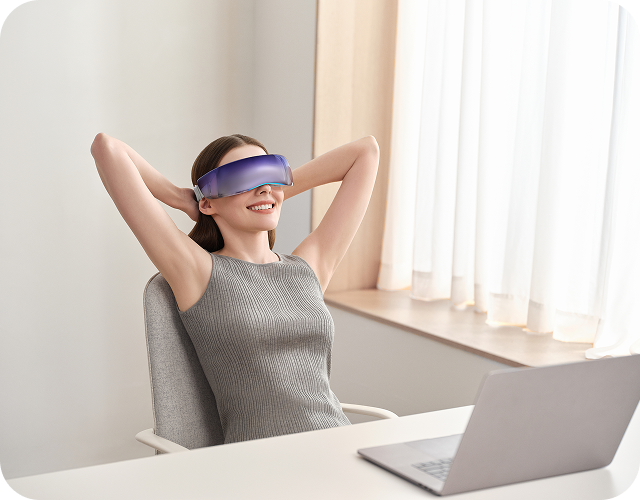 Uśmiechnięta kobieta w biurze relaksuje się w fotelu, używając futurystycznego masażera oczu SKG KS700