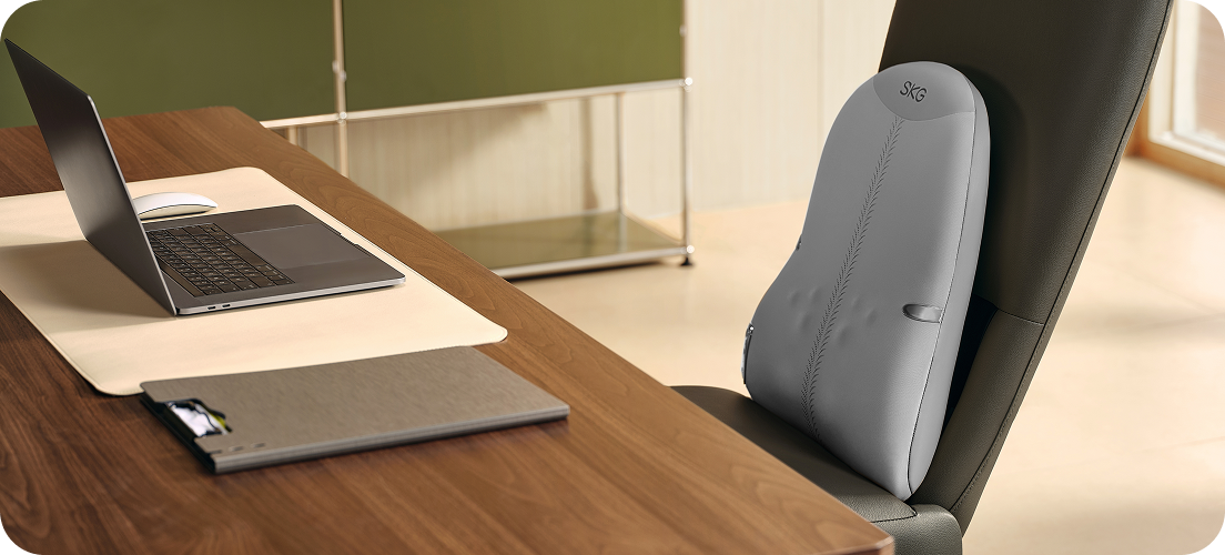 Szary masażer pleców SKG TS700 zamocowany na fotelu biurowym obok laptopa na nowoczesnym drewnianym biurku