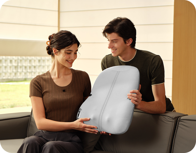 Uśmiechnięta para na kanapie, mężczyzna podaje kobiecie szary masażer pleców SKG TS700