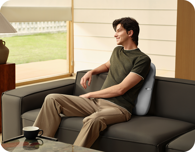 Uśmiechnięty mężczyzna siedzi na sofie, używając szarego masażera pleców SKG TS700 jako poduszki lędźwiowej