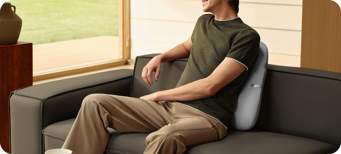 Uśmiechnięty mężczyzna siedzi na sofie, używając szarego masażera pleców SKG TS700 jako poduszki lędźwiowej