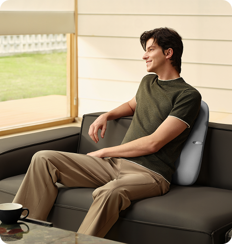 Uśmiechnięty mężczyzna siedzi na sofie, używając szarego masażera pleców SKG TS700 jako poduszki lędźwiowej