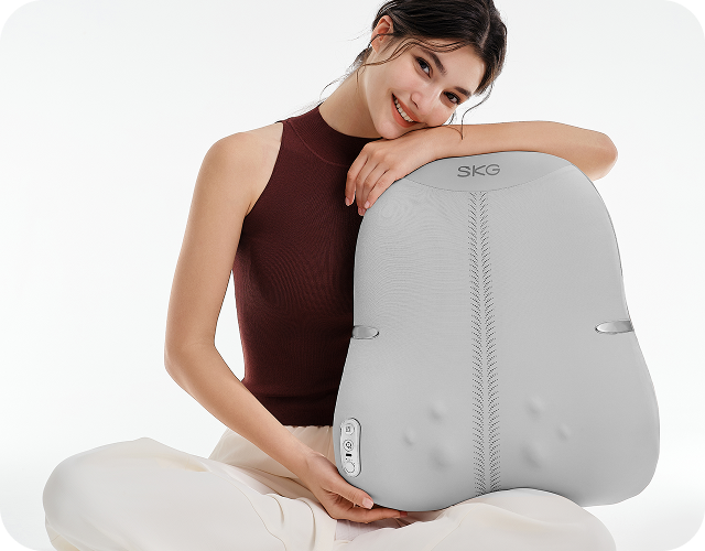 Uśmiechnięta kobieta w pozie lotosu trzyma duży, szary masażer pleców SKG TS700 na białym tle