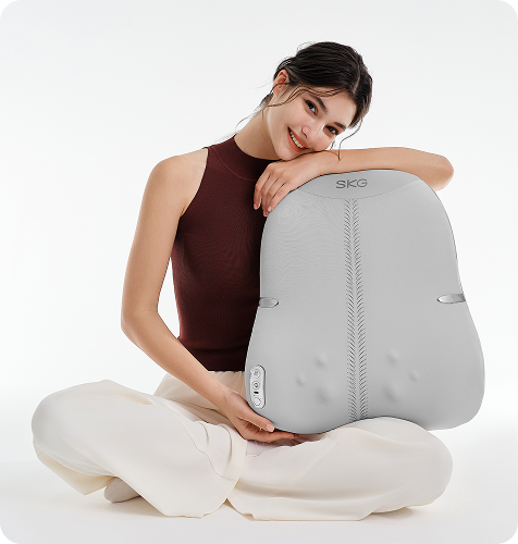 Uśmiechnięta kobieta w pozie lotosu trzyma duży, szary masażer pleców SKG TS700 na białym tle