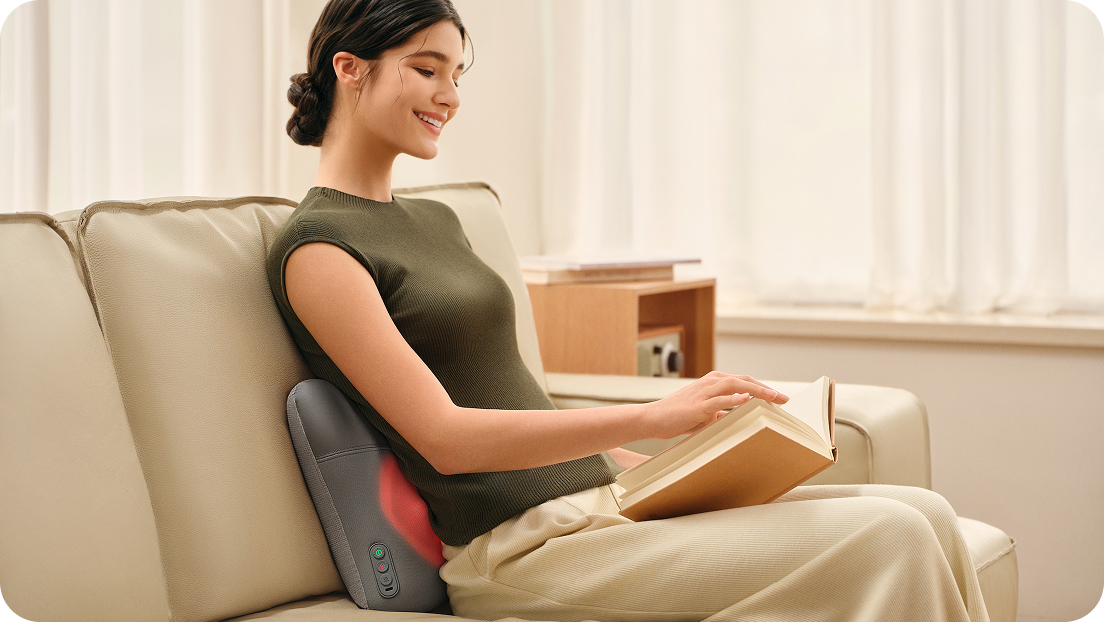 Uśmiechnięta kobieta czyta książkę, siedząc na kanapie i używając masażera pleców SKG VS500 z podgrzewaniem