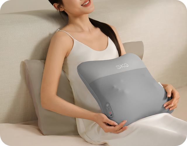 Kobieta relaksuje się na kanapie, używając masażera pleców SKG VS500 w kształcie poduszki na swoim brzuchu