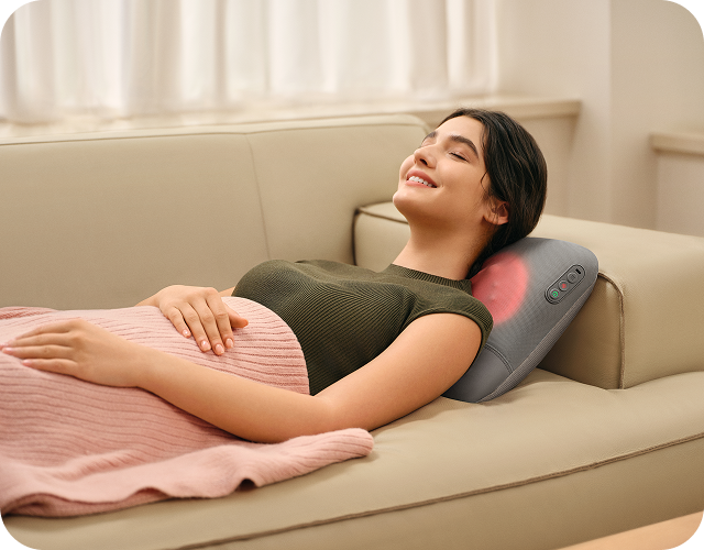 Uśmiechnięta kobieta leży na kanapie, używając masażera pleców SKG VS500 z funkcją podgrzewania szyi