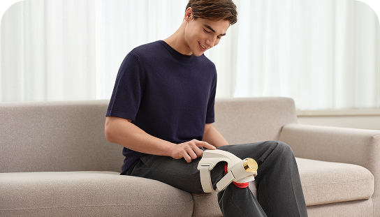 Mężczyzna siedzi na jasnej sofie i używa beżowo-złotego masażera SKG WS500 na kolanie