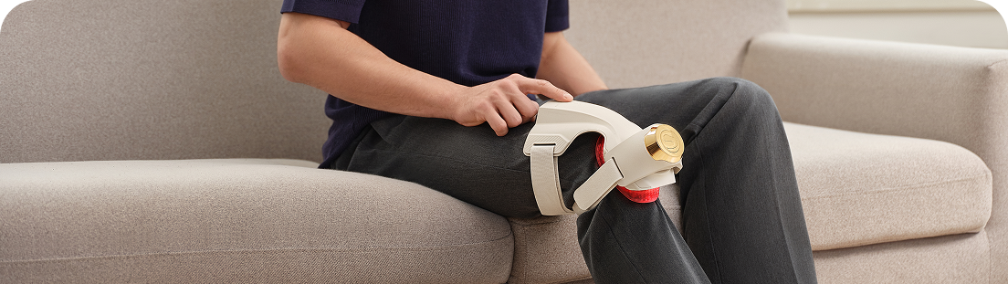 Mężczyzna siedzi na jasnej sofie i używa beżowo-złotego masażera SKG WS500 na kolanie