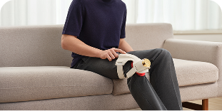 Mężczyzna siedzi na jasnej sofie i używa beżowo-złotego masażera SKG WS500 na kolanie