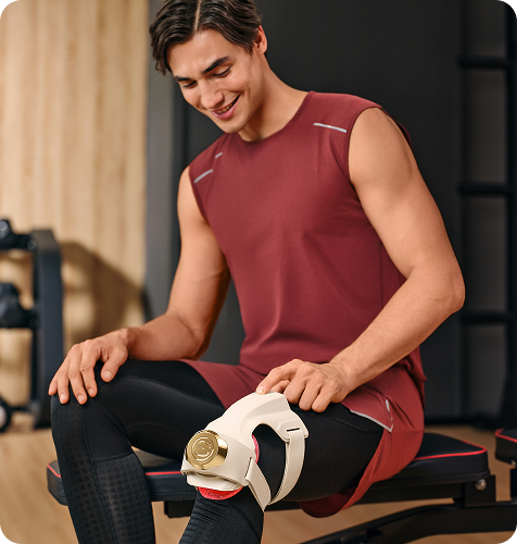 Uśmiechnięty mężczyzna siedzi na ławce do ćwiczeń i używa beżowo-złotego masażera SKG WS500 na kolanie